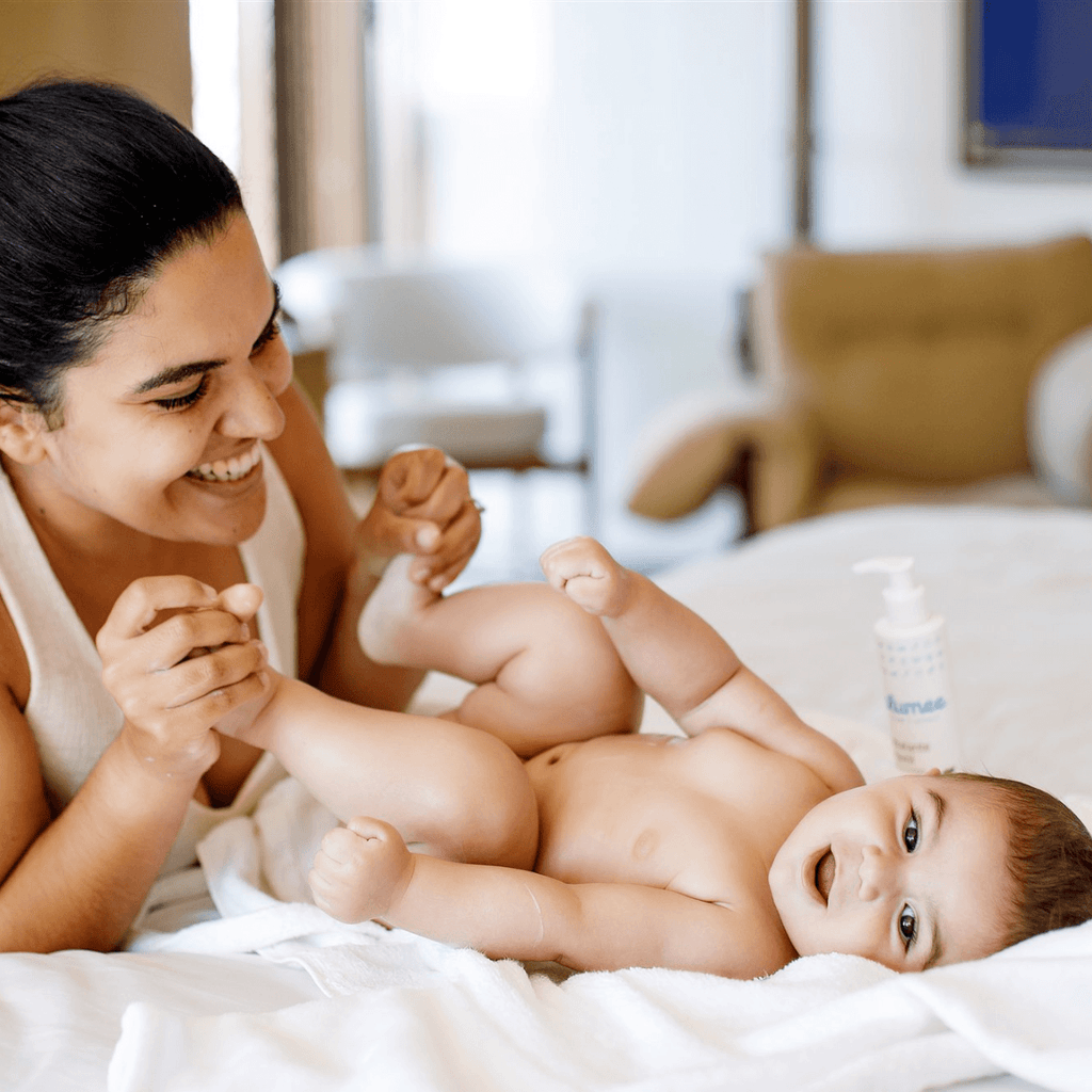 Mãe sorrindo brincando com os pés do bebê na cama, Hidratante Blumee ao fundo
