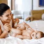 Mãe sorrindo brincando com os pés do bebê na cama, Hidratante Blumee ao fundo
