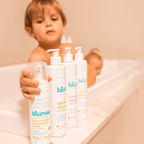 Criança no banho segurando produtos do Kit Blumee, com irmão ao fundo